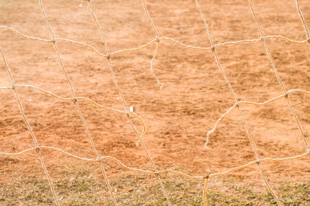 Textura com rede de futebol, luz do dia, grama, terra ao fundo.