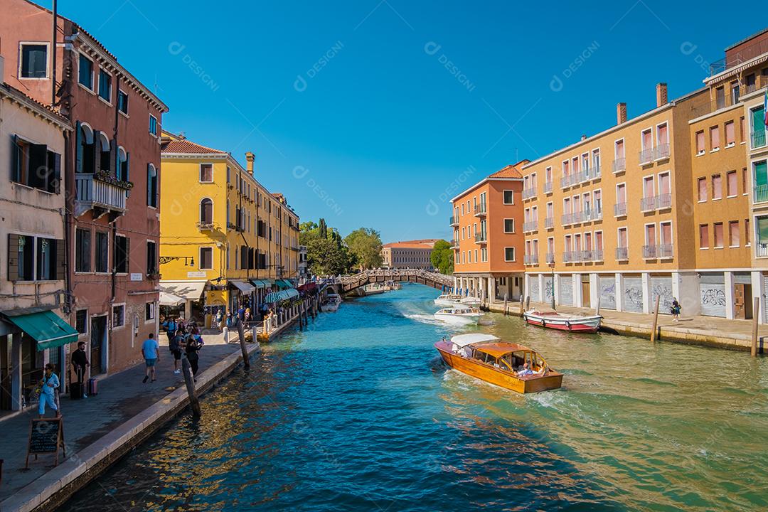 Vista de turistas andando na calçada ao lado do canal de Veneza com barco táxi e outros barcos passando.