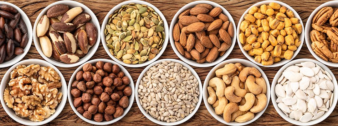 Sortidas de nozes e sementes em tigelas sobre mesa de madeira, formato panorâmico.