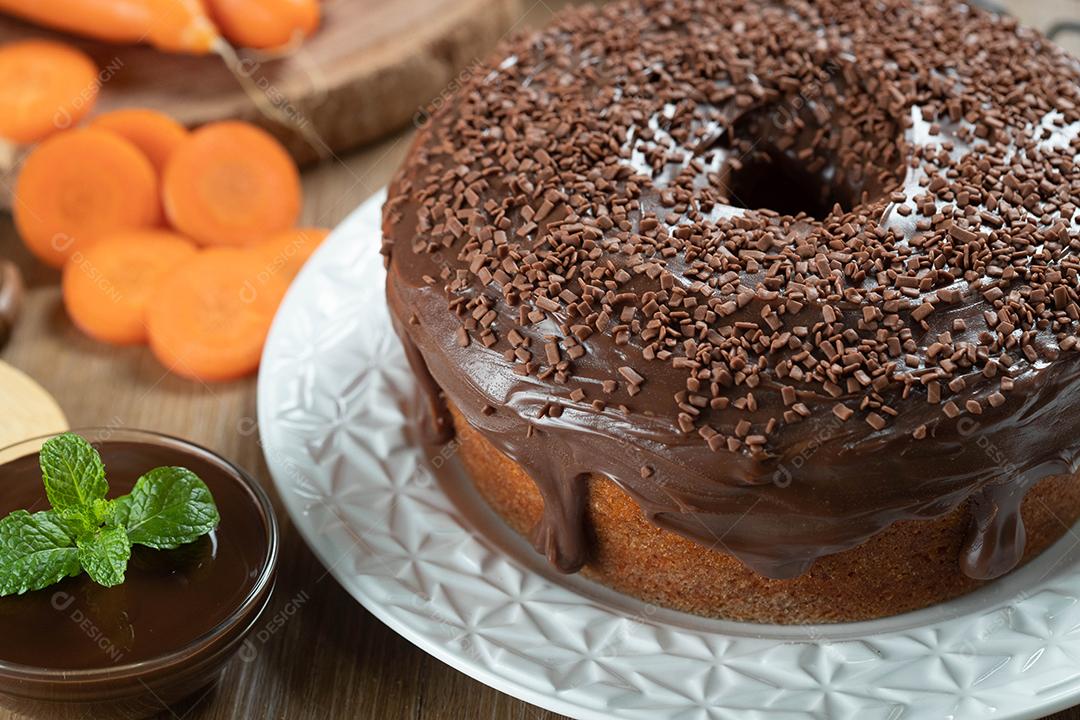 Bolo de cenoura brasileiro com cobertura de chocolate na mesa de madeira com cenouras ao fundo.