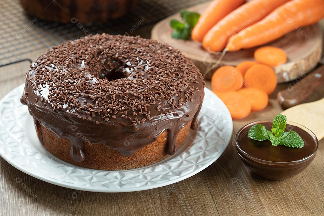 Brazilian carrot cake with chocolate icing on wooden table with carrots in the background.