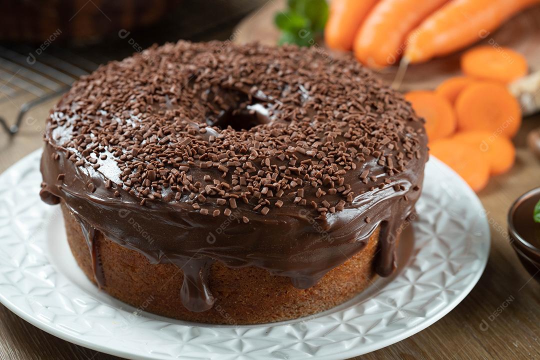 Bolo de cenoura brasileiro com cobertura de chocolate na mesa de madeira com cenouras ao fundo.