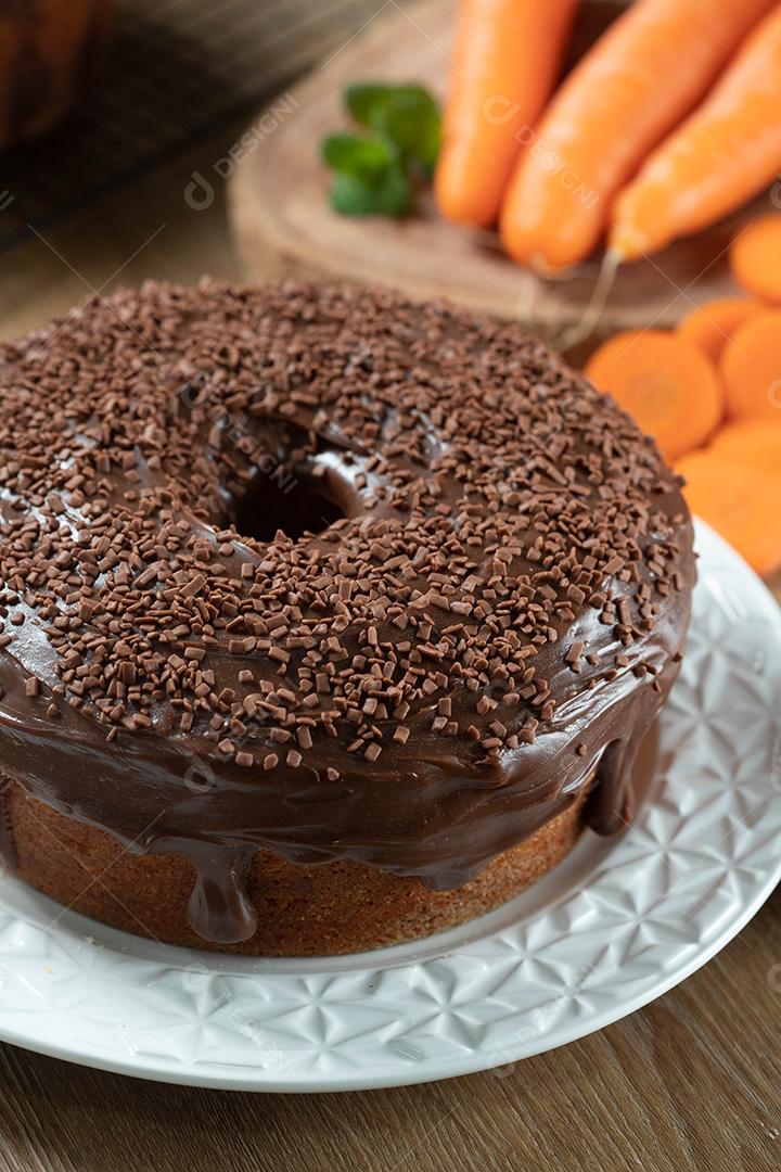 Bolo de cenoura brasileiro com cobertura de chocolate na mesa de madeira com cenouras ao fundo.