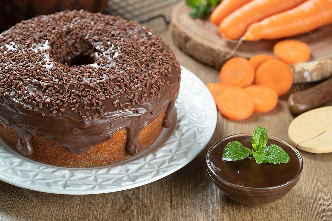 Brazilian carrot cake with chocolate icing on wooden table with carrots in the background.
