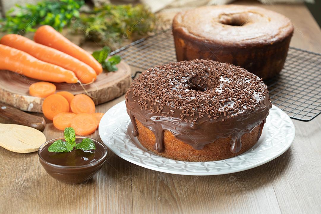Bolo de cenoura brasileiro com cobertura de chocolate na mesa de madeira com cenouras ao fundo.