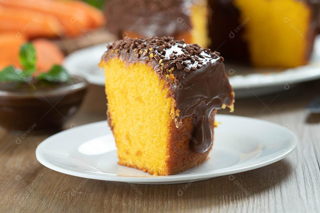 Feche o pedaço de bolo de cenoura brasileiro com cobertura de chocolate na mesa de madeira com cenouras ao fundo.