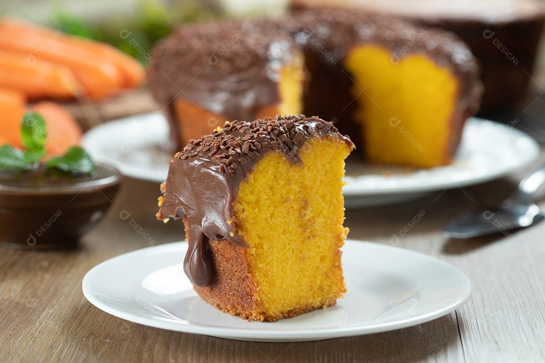 Feche o pedaço de bolo de cenoura brasileiro com cobertura de chocolate na mesa de madeira com cenouras ao fundo.