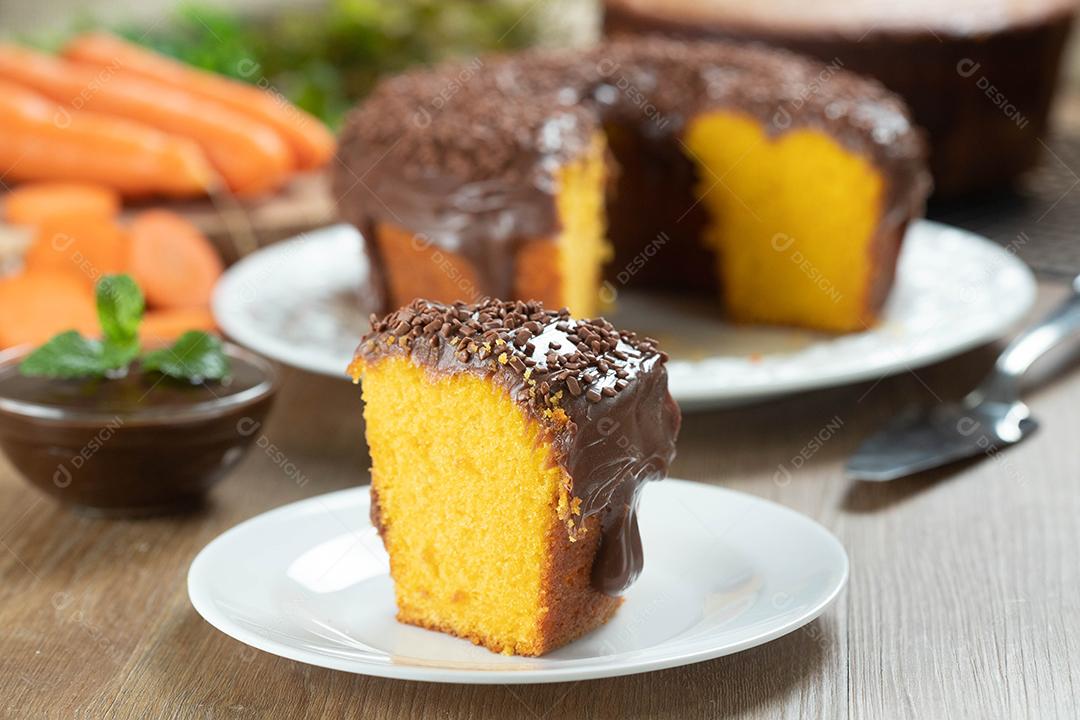 Close up piece of Brazilian carrot cake with chocolate icing on wooden table with carrots in the background.