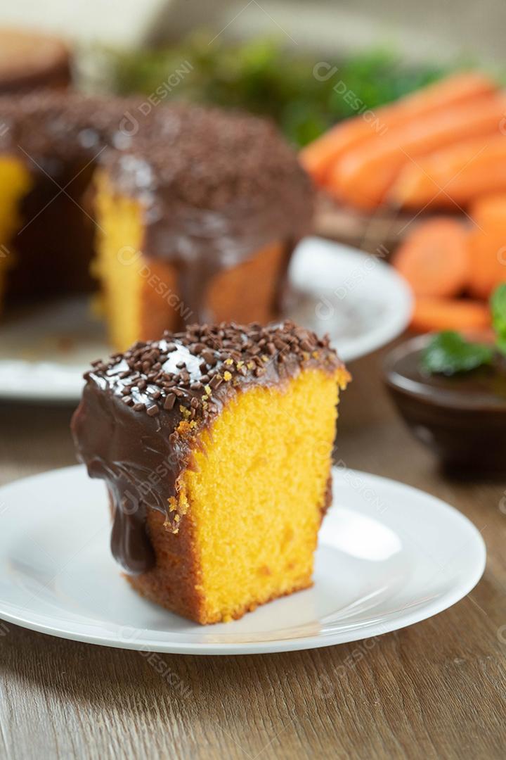 Feche o pedaço de bolo de cenoura brasileiro com cobertura de chocolate na mesa de madeira com cenouras ao fundo.