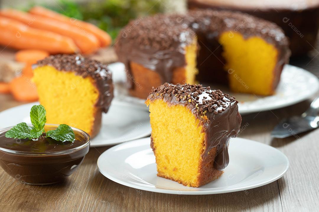 Feche o pedaço de bolo de cenoura brasileiro com cobertura de chocolate na mesa de madeira com cenouras ao fundo.