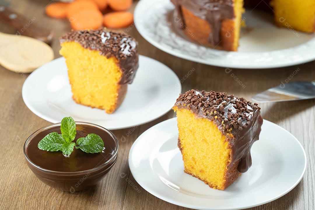 Feche o pedaço de bolo de cenoura brasileiro com cobertura de chocolate na mesa de madeira com cenouras ao fundo.