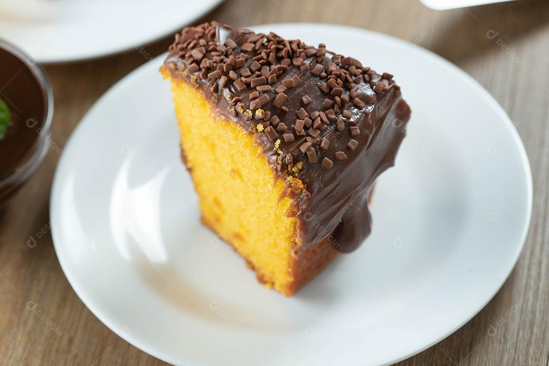Feche o pedaço de bolo de cenoura brasileiro com cobertura de chocolate na mesa de madeira com cenouras ao fundo.