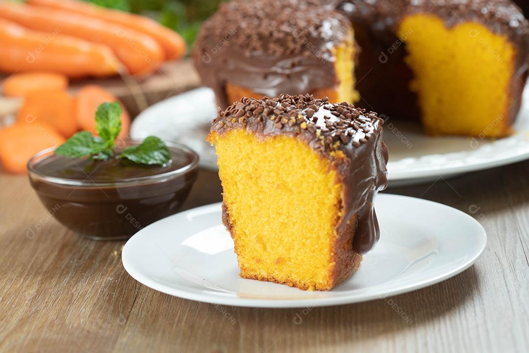 Feche o pedaço de bolo de cenoura brasileiro com cobertura de chocolate na mesa de madeira com cenouras ao fundo.