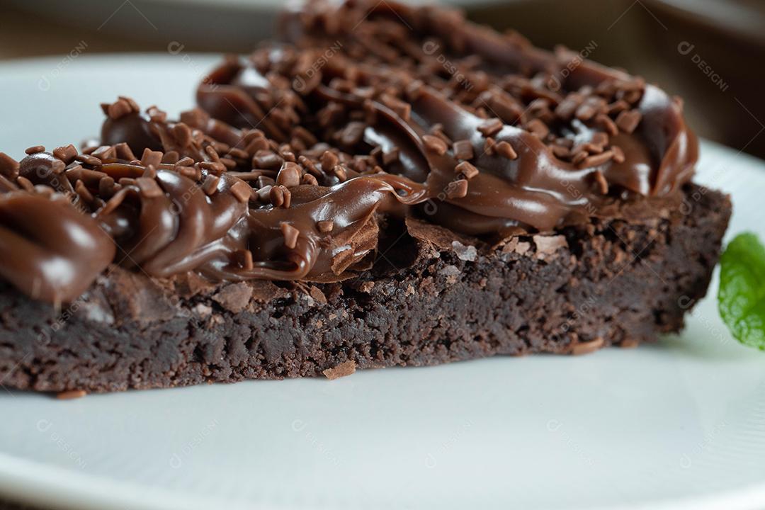 Fatia de brownie de chocolate com morango e cobertura de chocolate. Mesa de madeira com menta e lascas de chocolate ao fundo