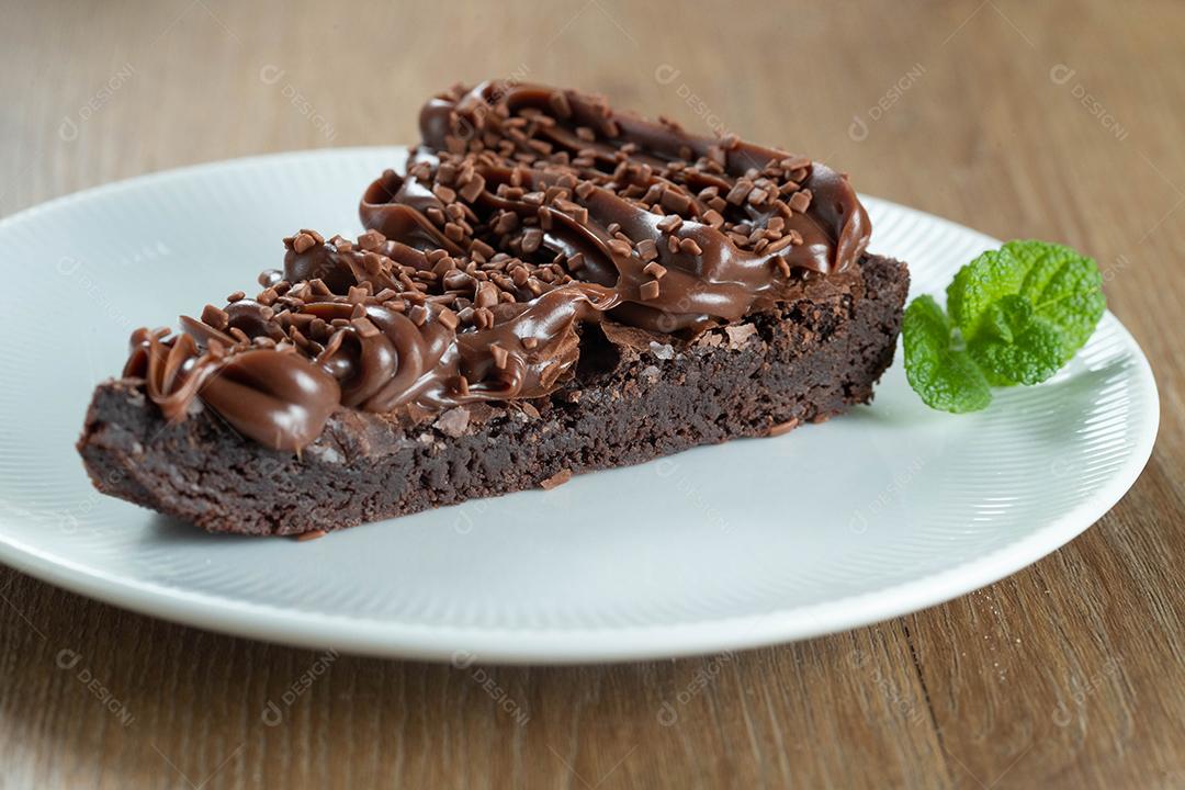 Fatia de brownie de chocolate com morango e cobertura de chocolate. Mesa de madeira com menta e lascas de chocolate ao fundo