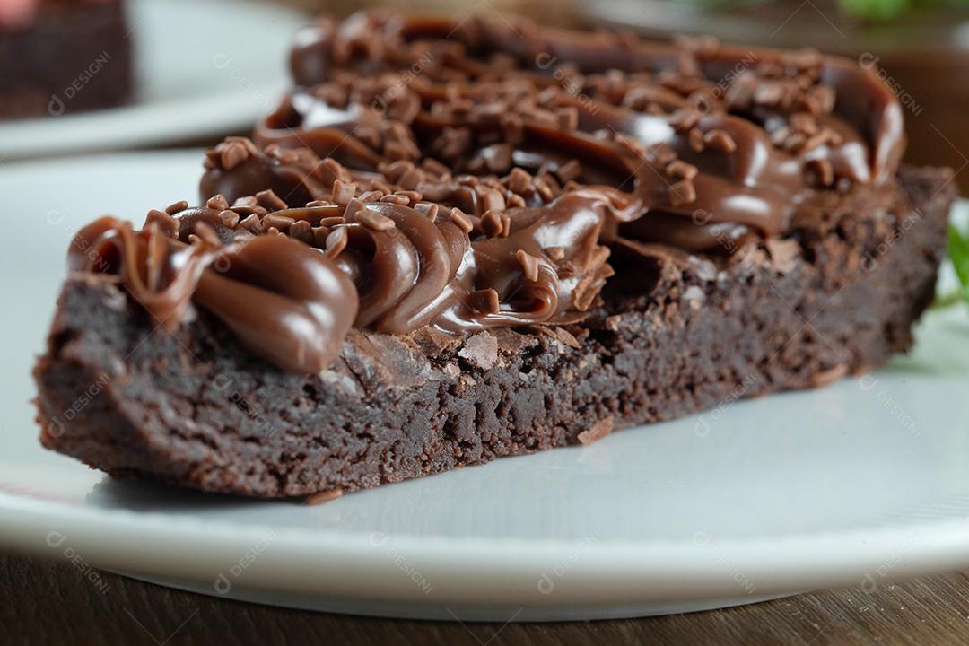 Fatia de brownie de chocolate com morango e cobertura de chocolate. Mesa de madeira com menta e lascas de chocolate ao fundo