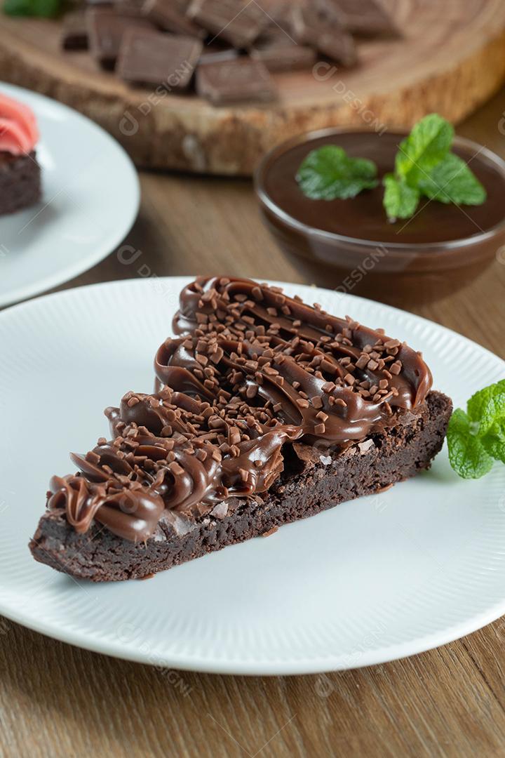 Fatia de brownie de chocolate com morango e cobertura de chocolate. Mesa de madeira com menta e lascas de chocolate ao fundo.