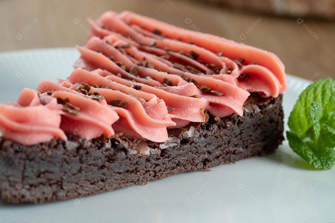 Fatia de brownie de chocolate com morango e cobertura de chocolate. Mesa de madeira com menta e lascas de chocolate ao fundo.