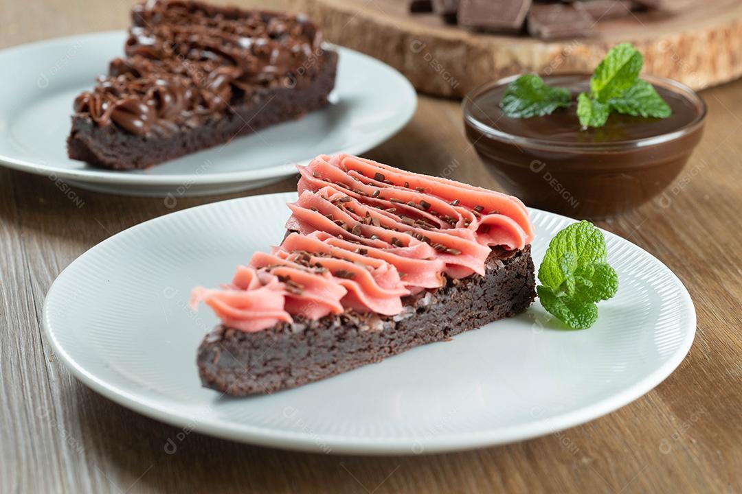 Fatia de brownie de chocolate com morango e cobertura de chocolate. Mesa de madeira com menta e lascas de chocolate ao fundo.