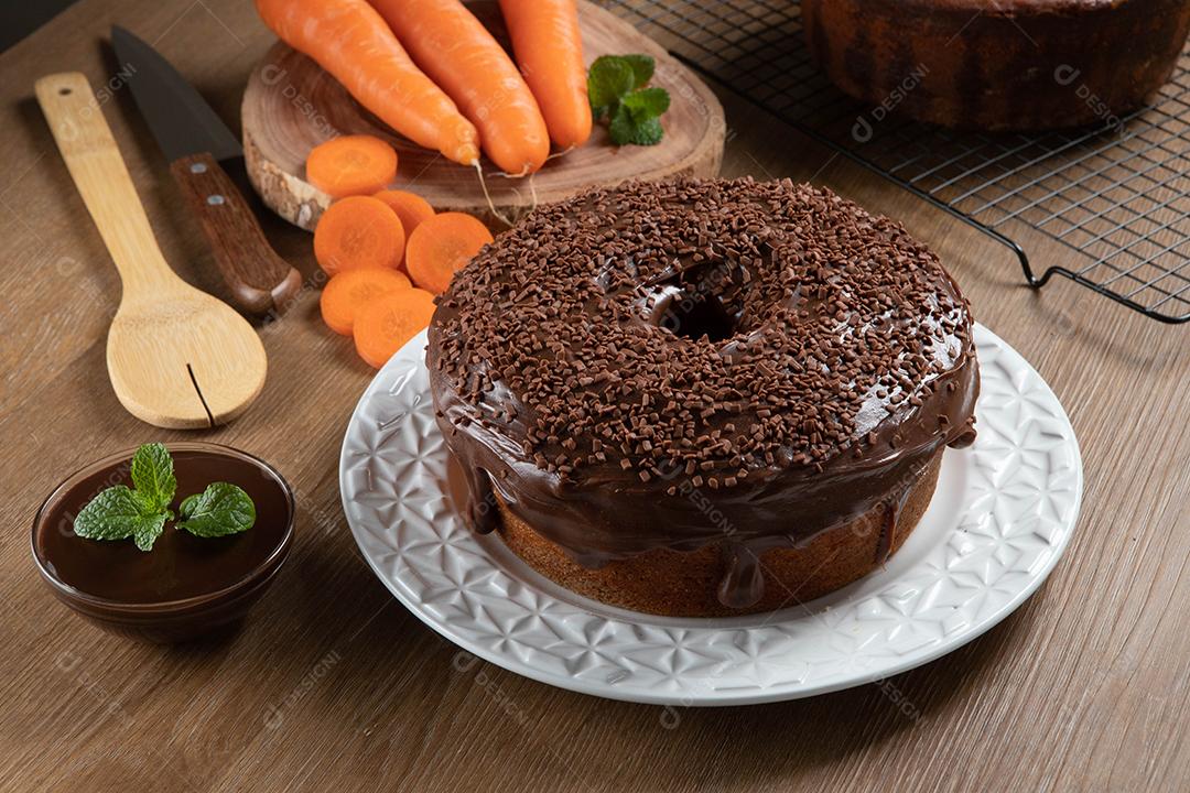 Bolo de cenoura brasileiro com cobertura de chocolate na mesa de madeira com cenouras ao fundo.