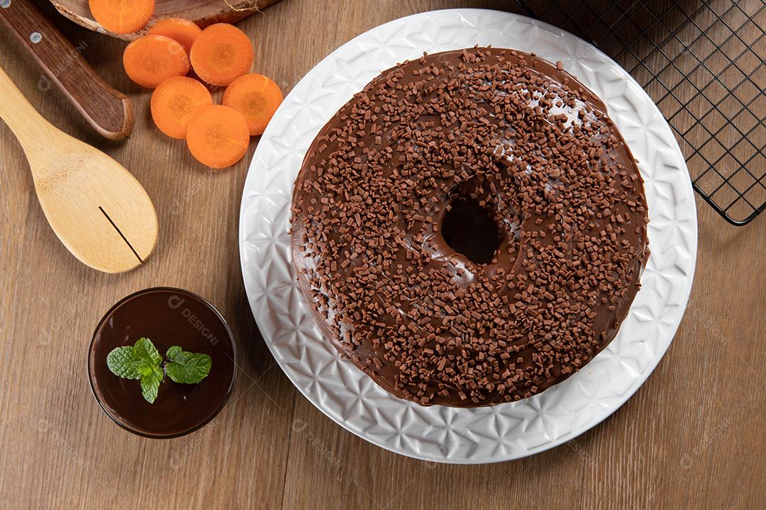 Bolo de cenoura brasileiro com cobertura de chocolate na mesa de madeira com cenouras ao fundo. Vista do topo