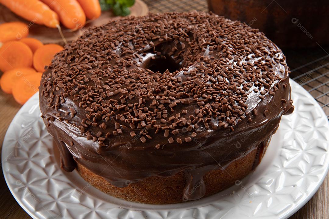 Bolo de cenoura brasileiro com cobertura de chocolate na mesa de madeira com cenouras ao fundo.