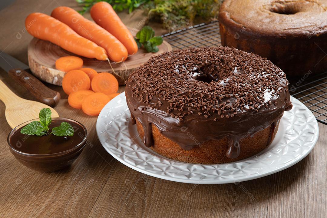 Bolo de cenoura brasileiro com cobertura de chocolate na mesa de madeira com cenouras ao fundo.