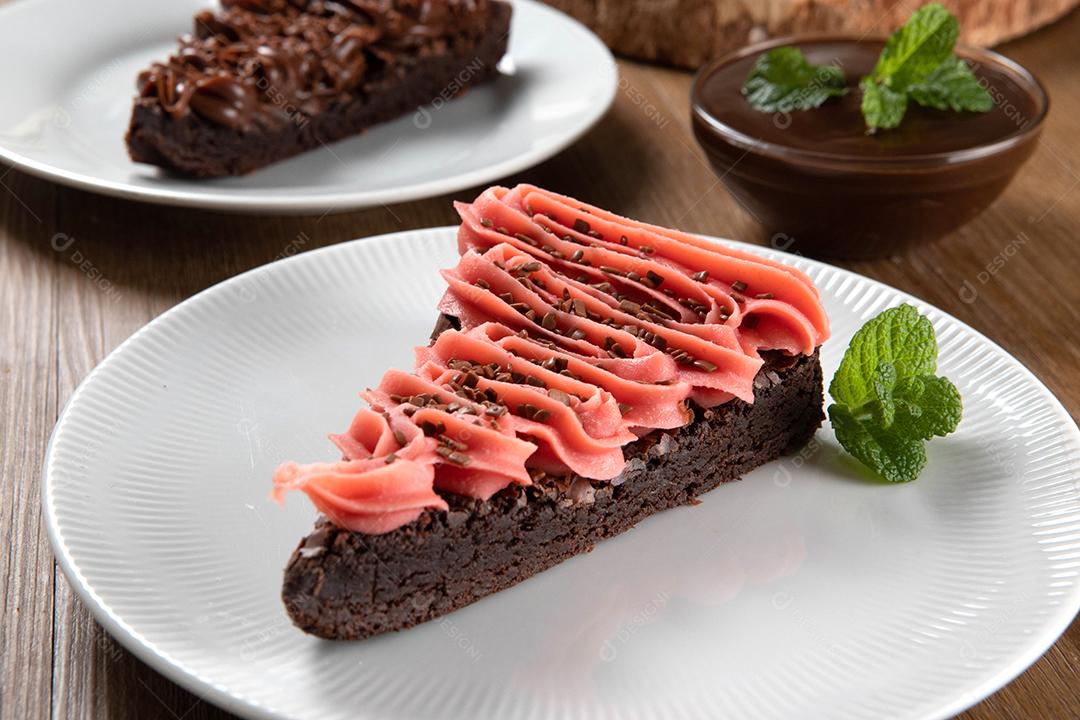 Fatia de brownie de chocolate com morango e cobertura de chocolate. Mesa de madeira com menta e lascas de chocolate ao fundo