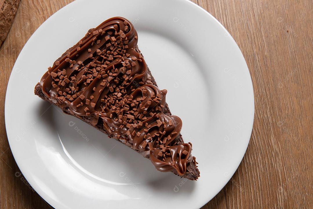 Fatia de brownie de chocolate com morango e cobertura de chocolate. Mesa de madeira com menta e lascas de chocolate ao fundo