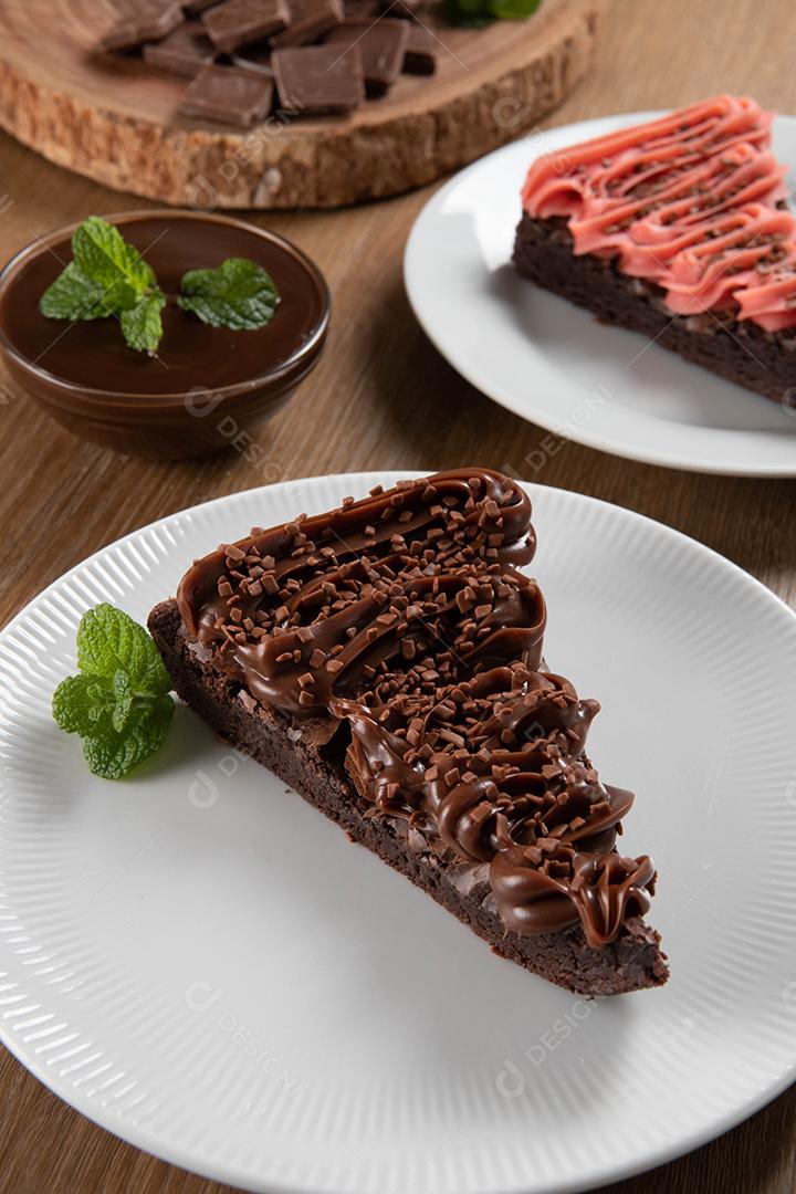 Fatia de brownie de chocolate com morango e cobertura de chocolate. Mesa de madeira com menta e lascas de chocolate ao fundo