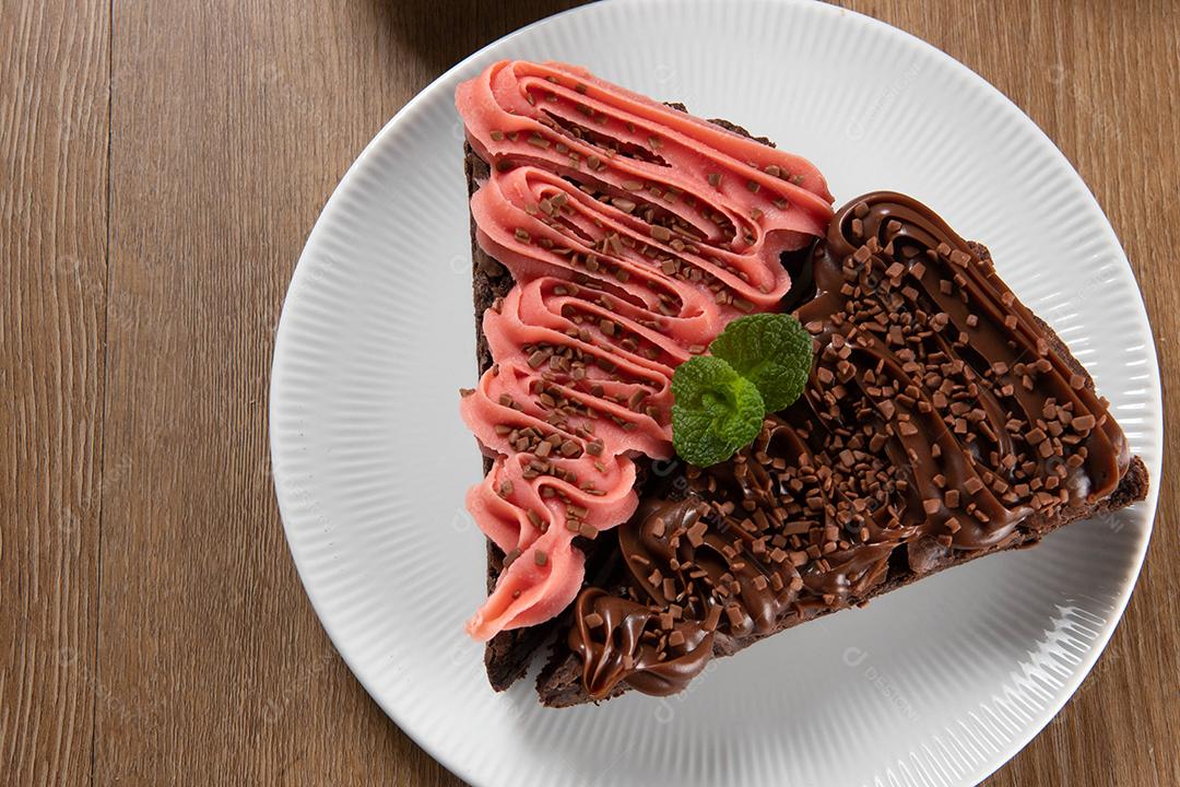 Fatia de brownie de chocolate com morango e cobertura de chocolate. Mesa de madeira com menta e lascas de chocolate ao fundo