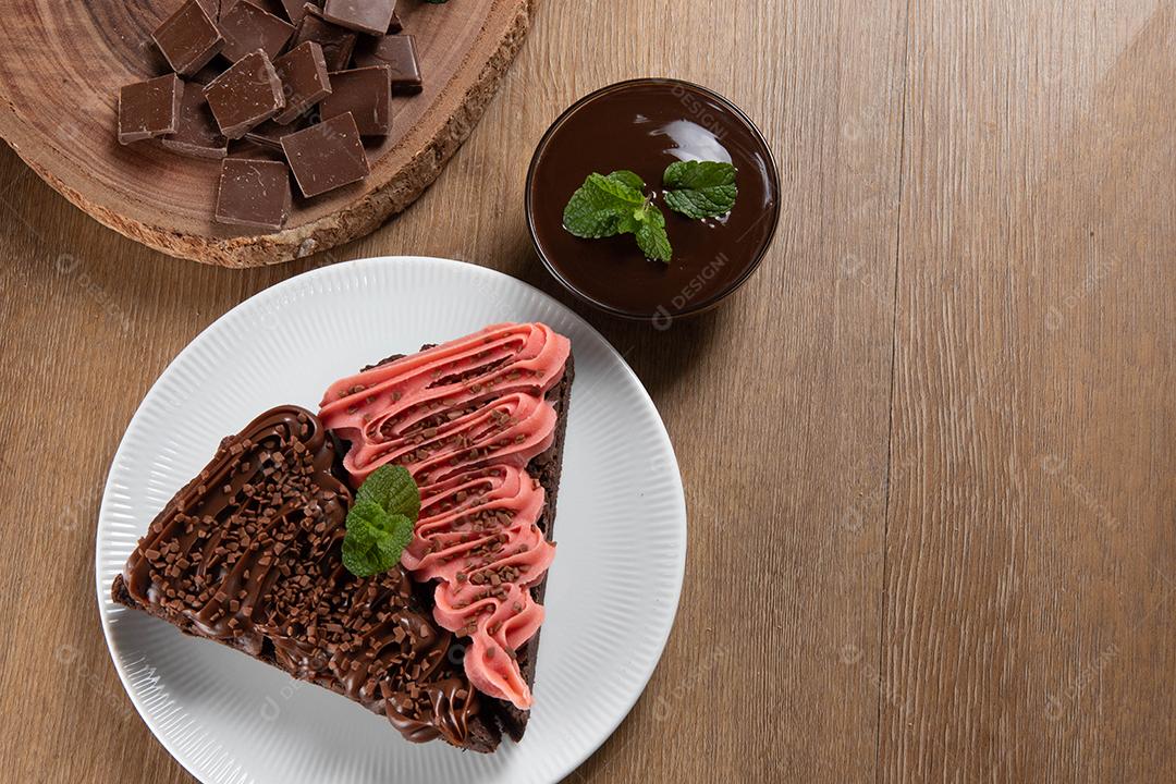 Fatia de brownie de chocolate com morango e cobertura de chocolate. Mesa de madeira com menta e lascas de chocolate ao fundo