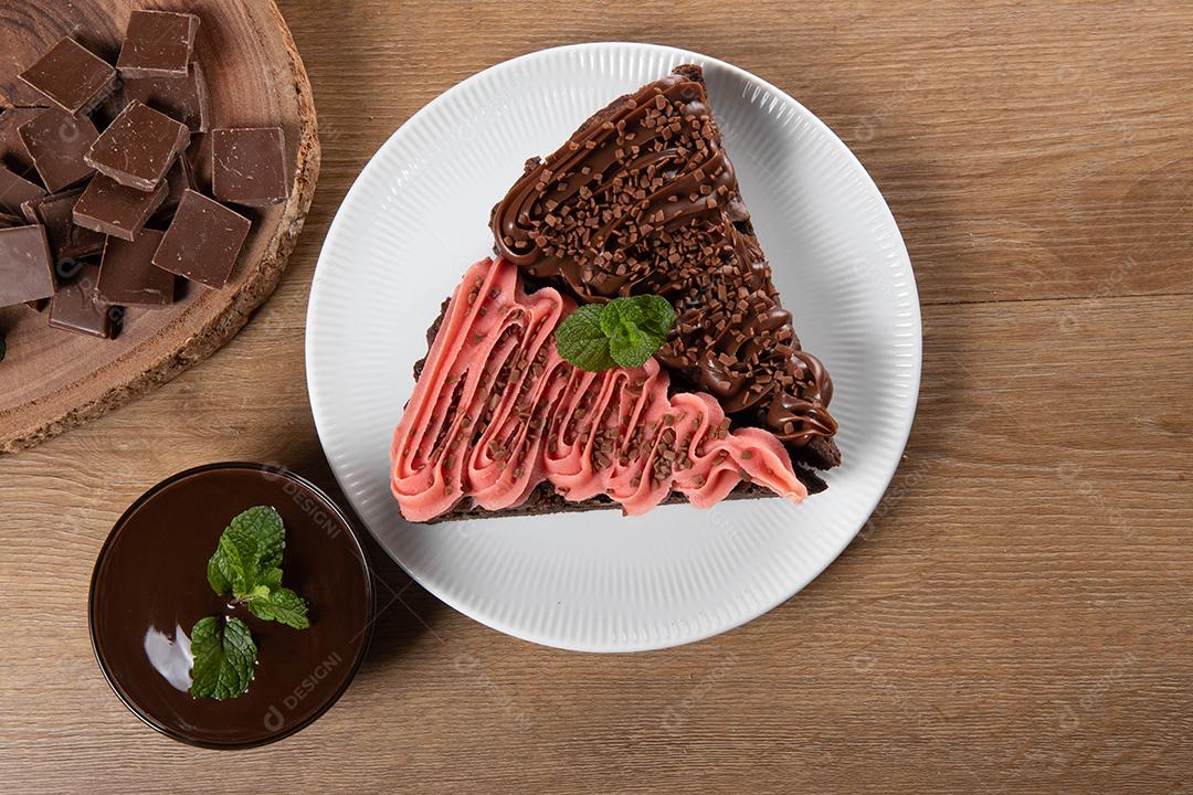 Fatia de brownie de chocolate com morango e cobertura de chocolate. Mesa de madeira com menta e lascas de chocolate ao fundo