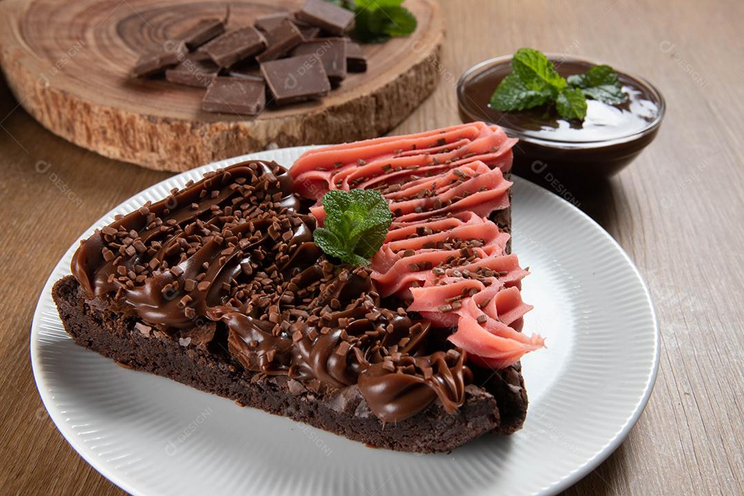Fatia de brownie de chocolate com morango e cobertura de chocolate. Mesa de madeira com menta e lascas de chocolate ao fundo