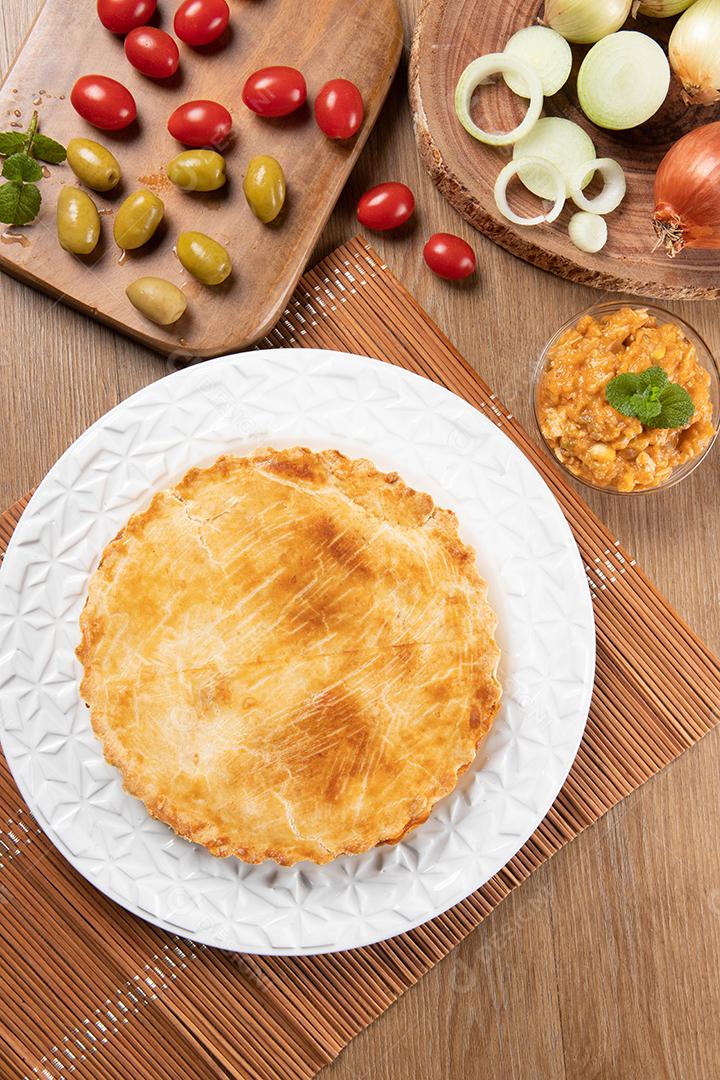 Torta de frango com requeijão, tomate, cebola e azeitonas na mesa de madeira e chapa branca. Vista do topo.