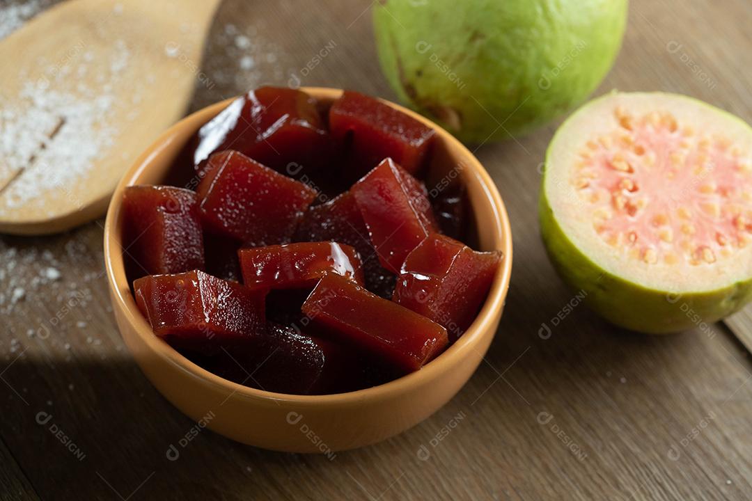 lata amarela com geléia de goiaba cortada em cubos com goiabas ao fundo na mesa de madeira.