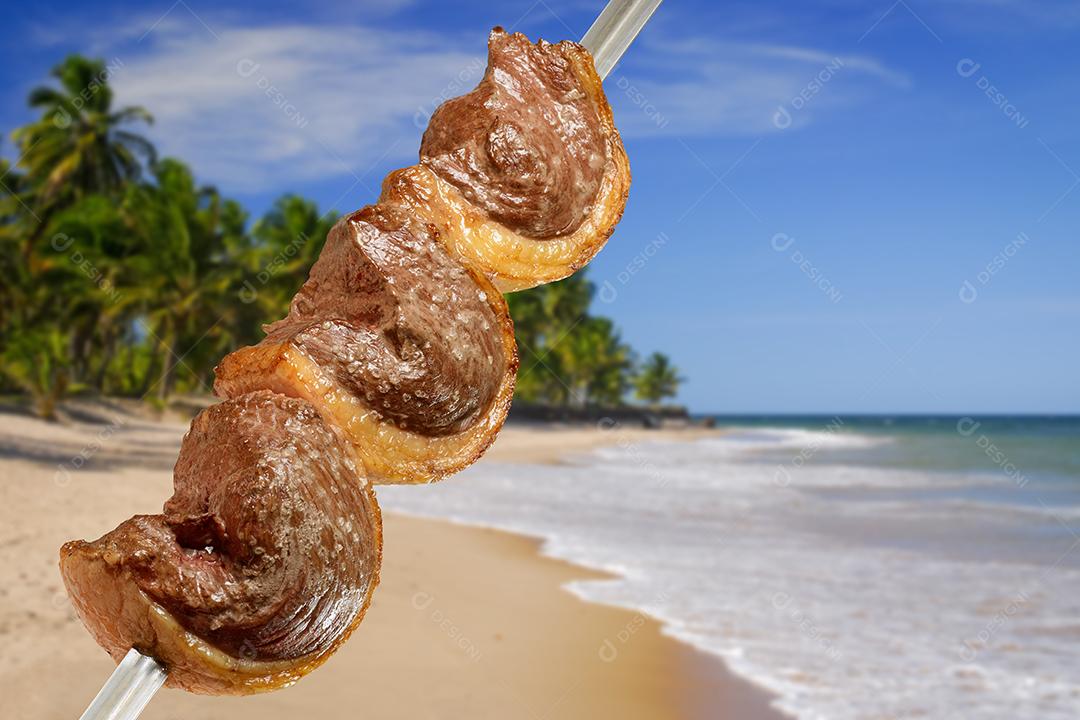 Picanha de churrasco grelhada no espeto com cena de praia brasileira ao fundo.