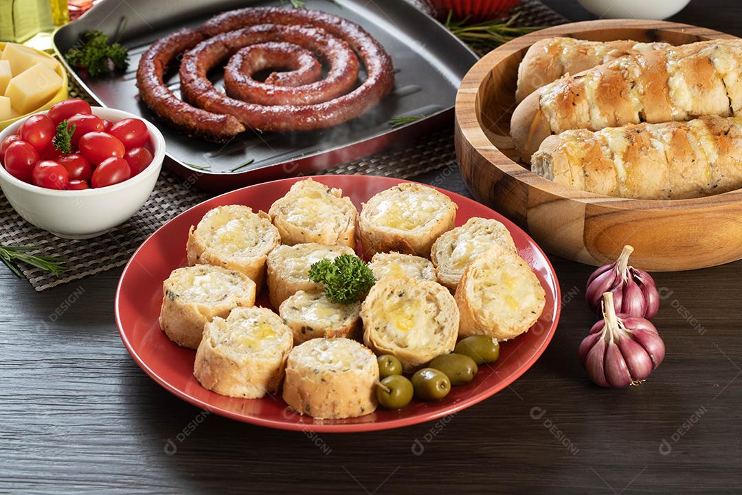 Pão de alho em um prato vermelho na mesa de churrasco com salsicha, queijo, alecrim, azeitonas e tomate cereja