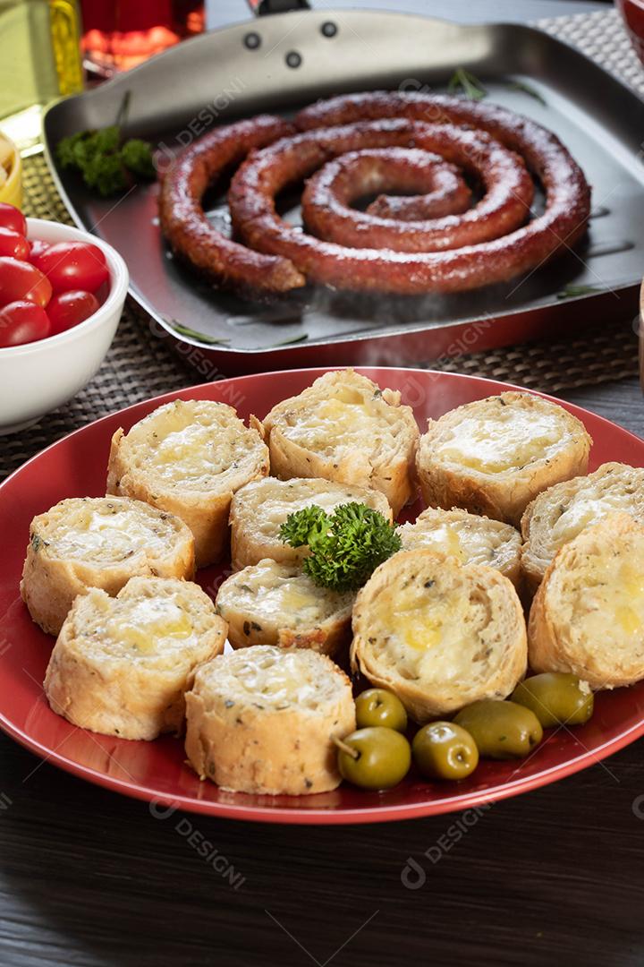 Pão de alho em um prato vermelho na mesa de churrasco com salsicha, queijo, alecrim, azeitonas e tomate cereja