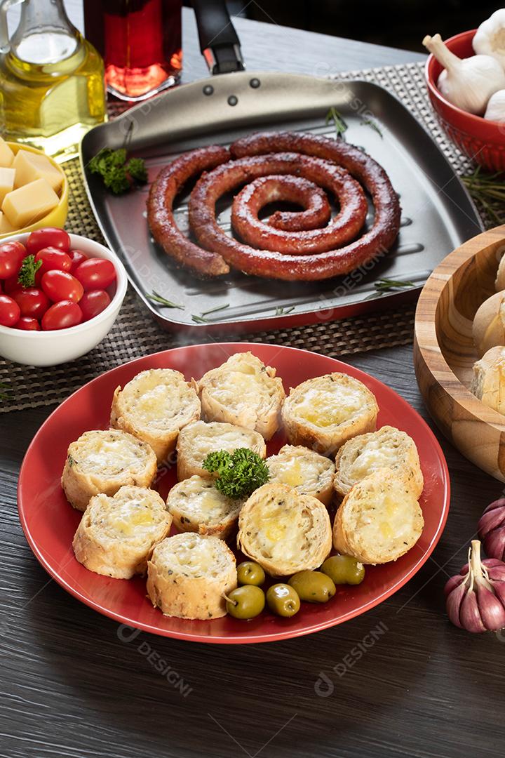 Pão de alho em um prato vermelho na mesa de churrasco com salsicha, queijo, alecrim, azeitonas e tomate cereja