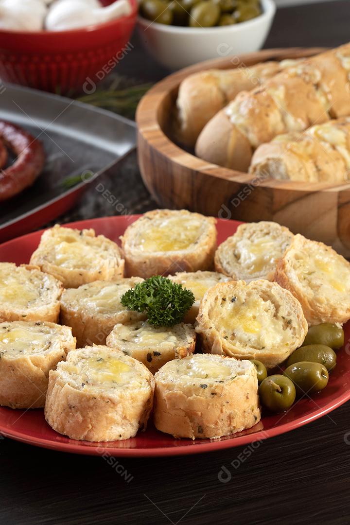 Pão de alho em um prato vermelho na mesa de churrasco com salsicha, queijo, alecrim, azeitonas e tomate cereja