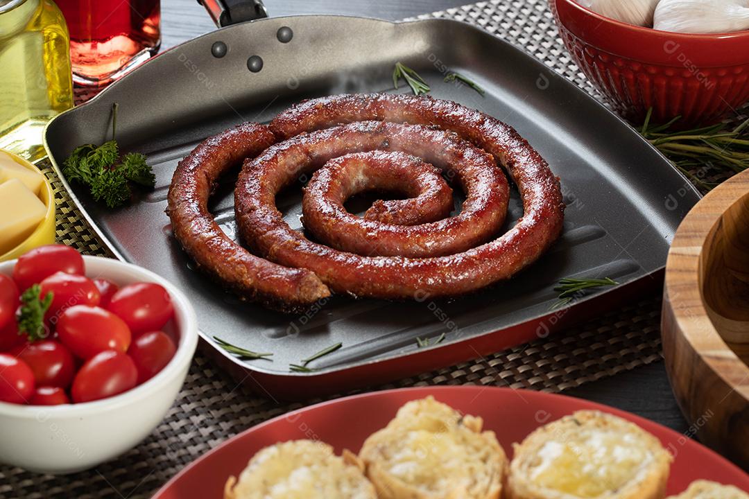 Pão de salsicha e alho em um prato vermelho na mesa de churrasco com aperitivos, queijo, alecrim, azeitonas e tomate cereja