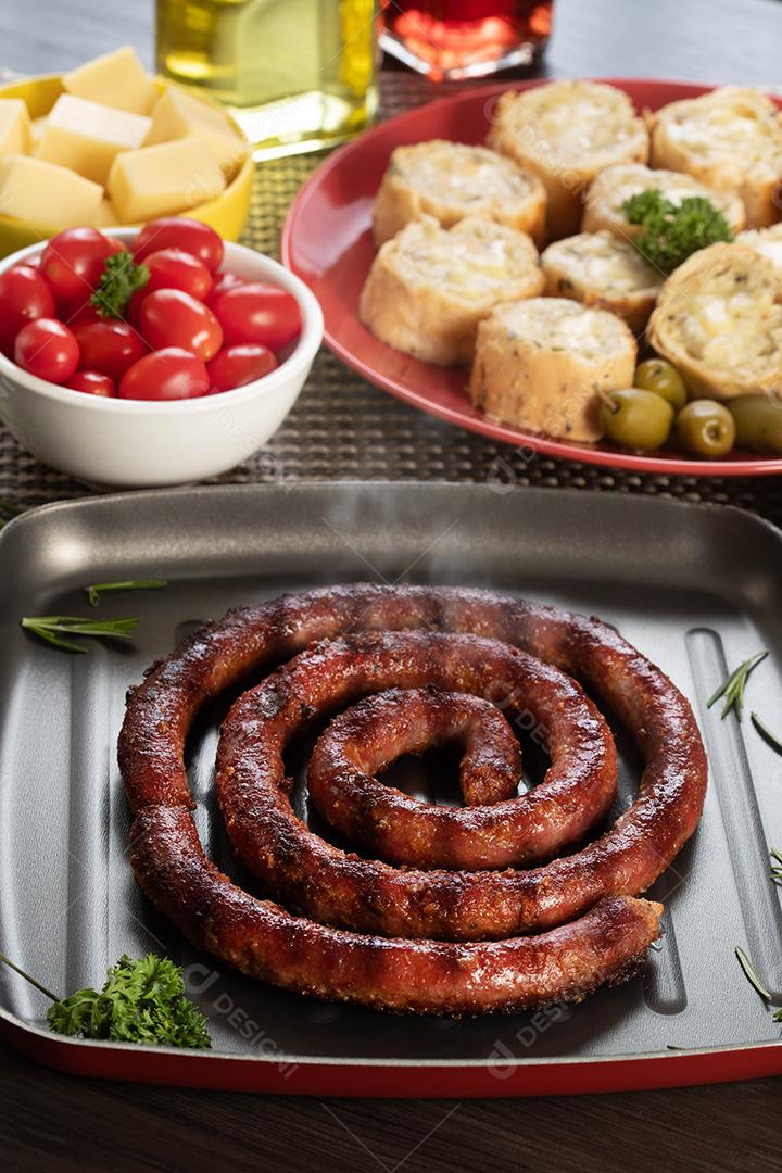 Pão de salsicha e alho em um prato vermelho na mesa de churrasco com aperitivos, queijo, alecrim, azeitonas e tomate cereja