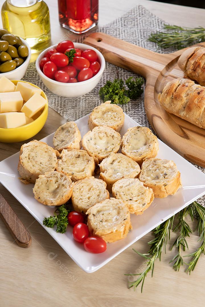 Pão de alho em prato quadrado branco em cima da mesa com queijo, alecrim, azeitonas e tomate cereja