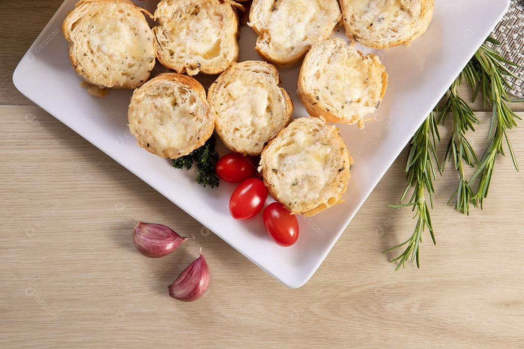 Pão de alho em prato quadrado branco em cima da mesa com queijo, alecrim, azeitonas e tomate cereja