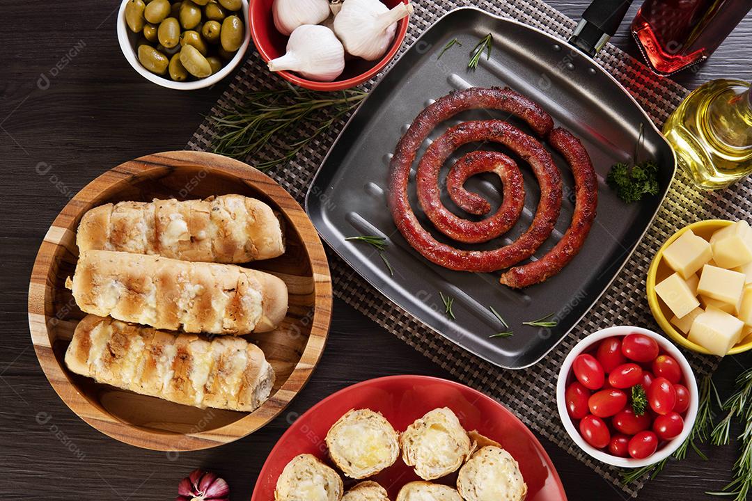 Pão de salsicha e alho em um prato vermelho na mesa de churrasco com aperitivos, queijo, alecrim, azeitonas e tomate cereja