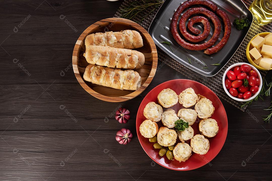 Pão de alho em um prato vermelho na mesa de churrasco com salsicha, queijo, alecrim, azeitonas e tomate cereja