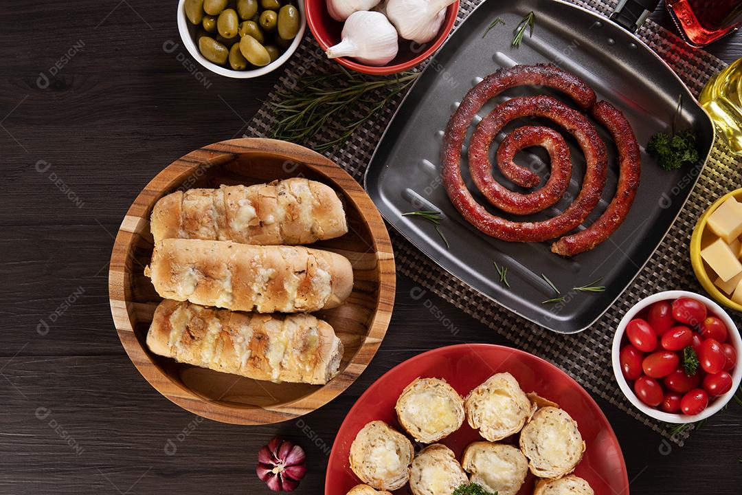 Pão de alho em um prato vermelho na mesa de churrasco com salsicha, queijo, alecrim, azeitonas e tomate cereja
