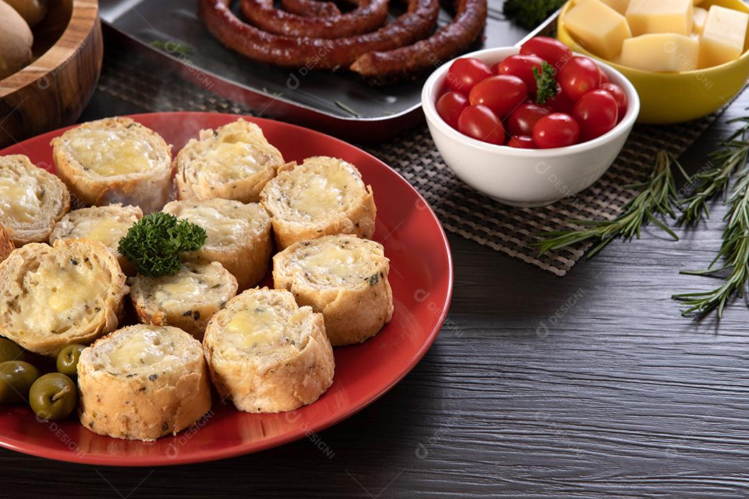 Pão de alho em um prato vermelho na mesa de churrasco com salsicha, queijo, alecrim, azeitonas e tomate cereja