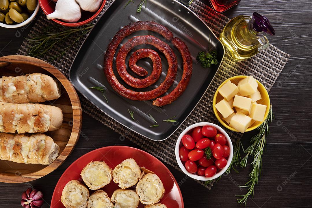 Pão de salsicha e alho em um prato vermelho na mesa de churrasco com aperitivos, queijo, alecrim, azeitonas e tomate cereja.